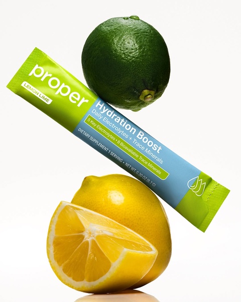 A high-quality product hero shot featuring a hydration electrolyte stick hovering between a whole lime and a cut lemon to emphasize fresh ingredients.